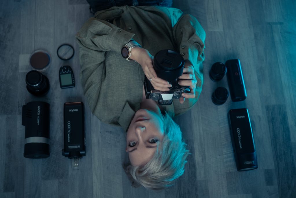 Das Bild zeigt eine Frau mit blonden Haaren und einer Kamera in der Hand. Bas Bild ist über Kopf, man sieht weitere Fotoausrüstung auf dem Boden liegen